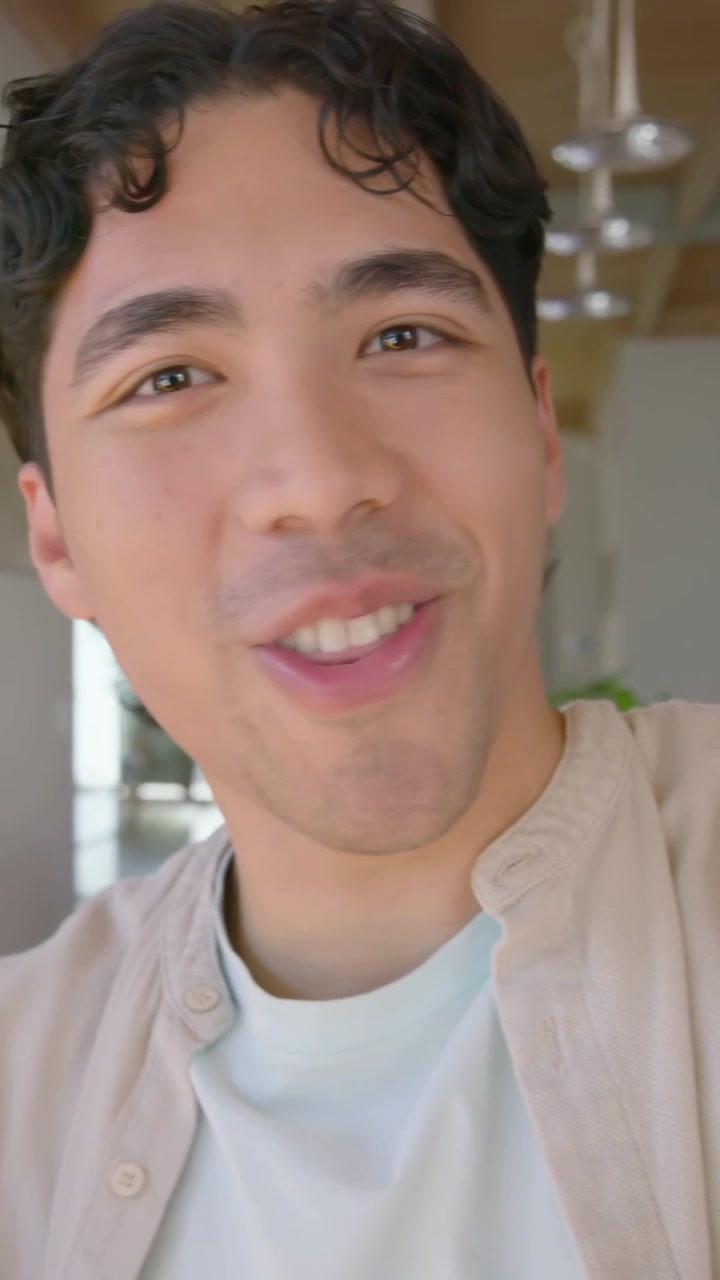Video verticale: Uomo in camicia beige preme "registra" e filma un discorso, sorridendo a casa con il telefono.