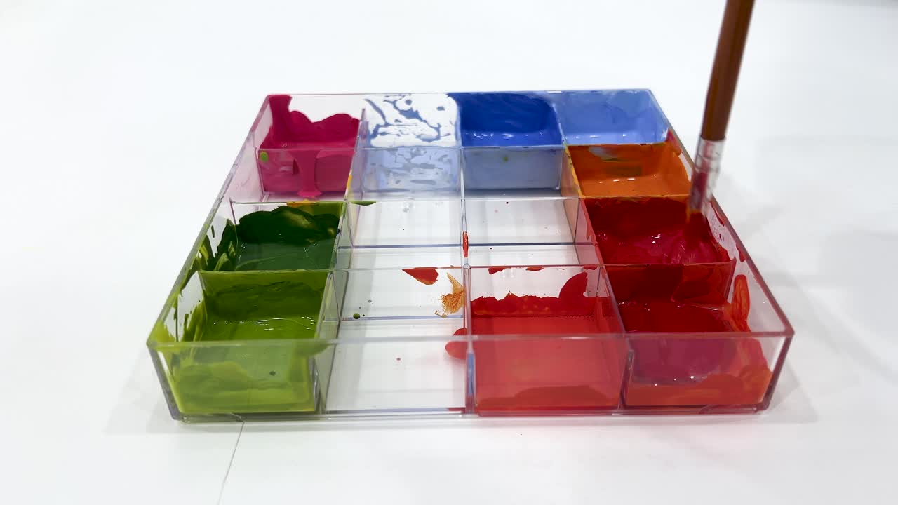 A hand uses a paintbrush to mix vibrant poster paints in a transparent tray on a white surface, under bright, even lighting with a static overhead camera