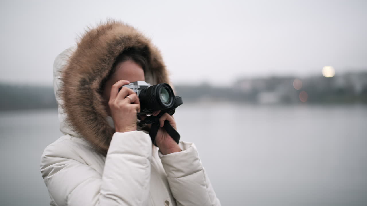 A woman in a warm coat stands by the water, using a camera to take photos of the serene landscape during a cool, overcast day. The quiet atmosphere enhances the moment