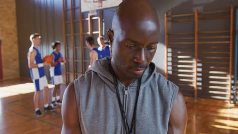 Portrait-of-african-american-male-basketball-coach-holding-clipboard-with-his-team-in-background