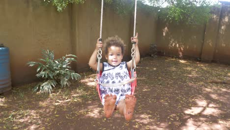 Una-Niña-Feliz-De-Raza-Mixta-Columpiándose-En-Un-Columpio-De-árbol-En-Un-Patio-Trasero-Mostrando-Sus-Pies-Sucios