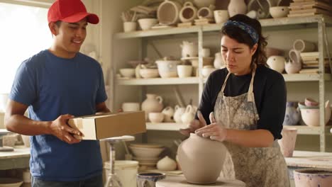 two pottery students interact in a bright studio. a young man hands a carefully wrapped ceramic piece to a young woman, showcasing their craftsmanship and teamwork with clay.