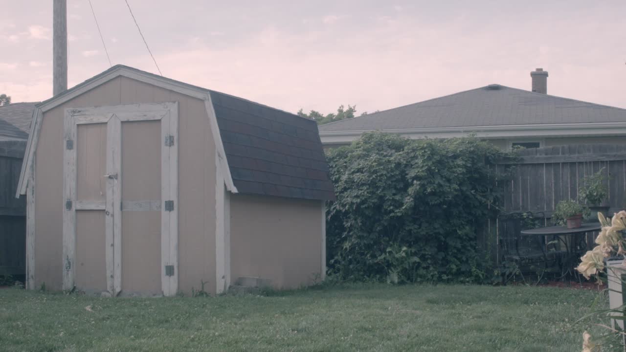 A timelapse shot in a suburban backyard during overcast conditions in Kenosha, Wisconsin. Features a beige barn-shed in the foreground, a hops plant behind it and a rotten out wooden fence.