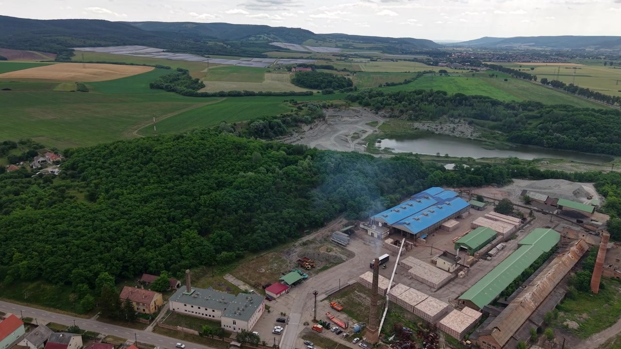 An aerial perspective captures an industrial facility with a smoking chimney situated near a rural village and vast green fields.