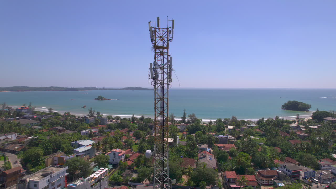 Fly around a telecommunications tower over the village. Aerial footage of telecommunications phone signal tower on tropical island.Flying around a phone signal tower with the ocean in the background
