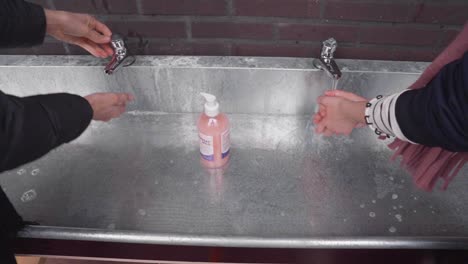 two girls washing hands with soap and water in metallic sink outside