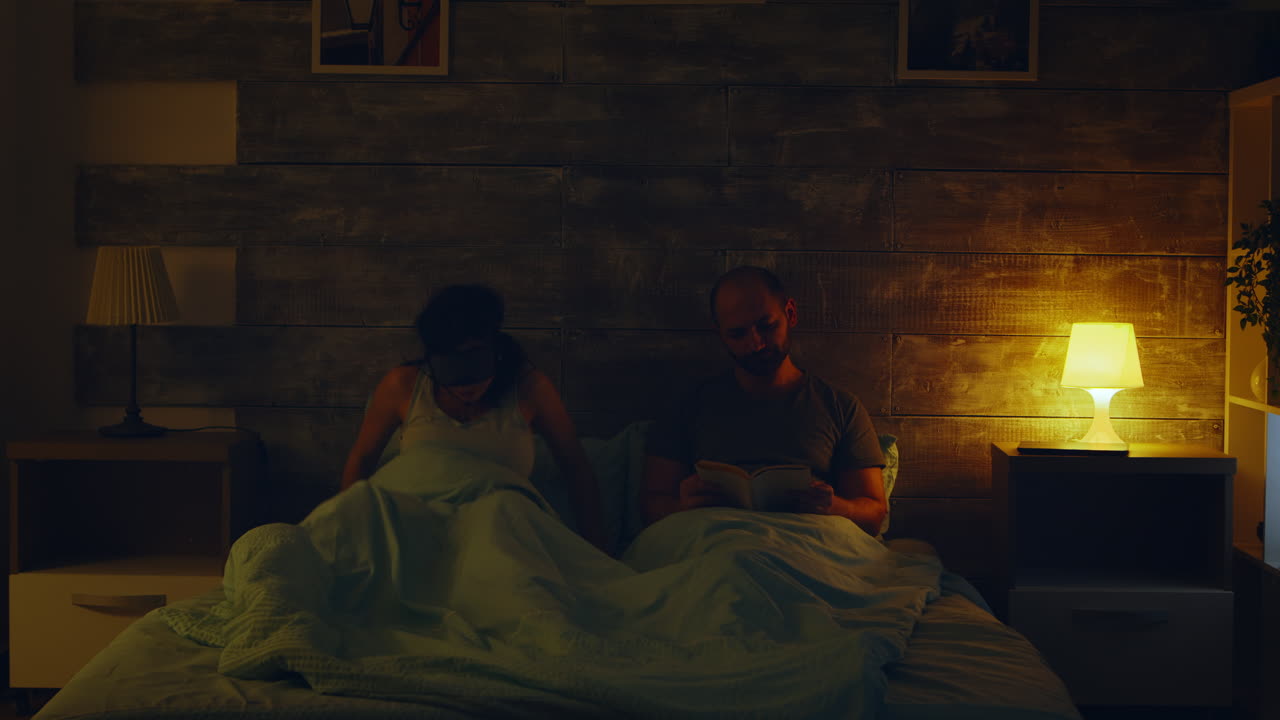 Young man reading a book while his wife is turning the lights off