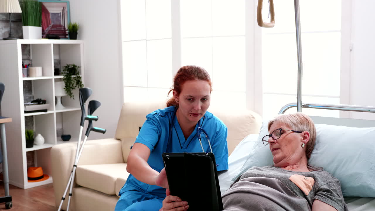 Old woman lying in bed learning from female doctor to use tablet computer