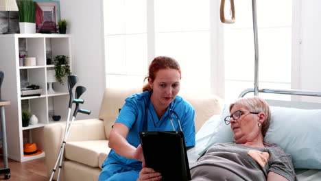 old woman lying in bed learning from female doctor to use tablet computer