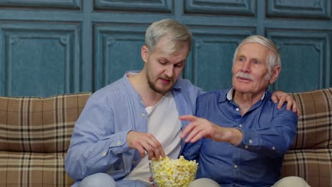 Älterer-Familienvater-Und-Erwachsener-Sohn-Sitzen-Auf-Dem-Sofa-Und-Schauen-Sich-Sportspiele-Im-Fernsehen-An-Und-Essen-Popcorn