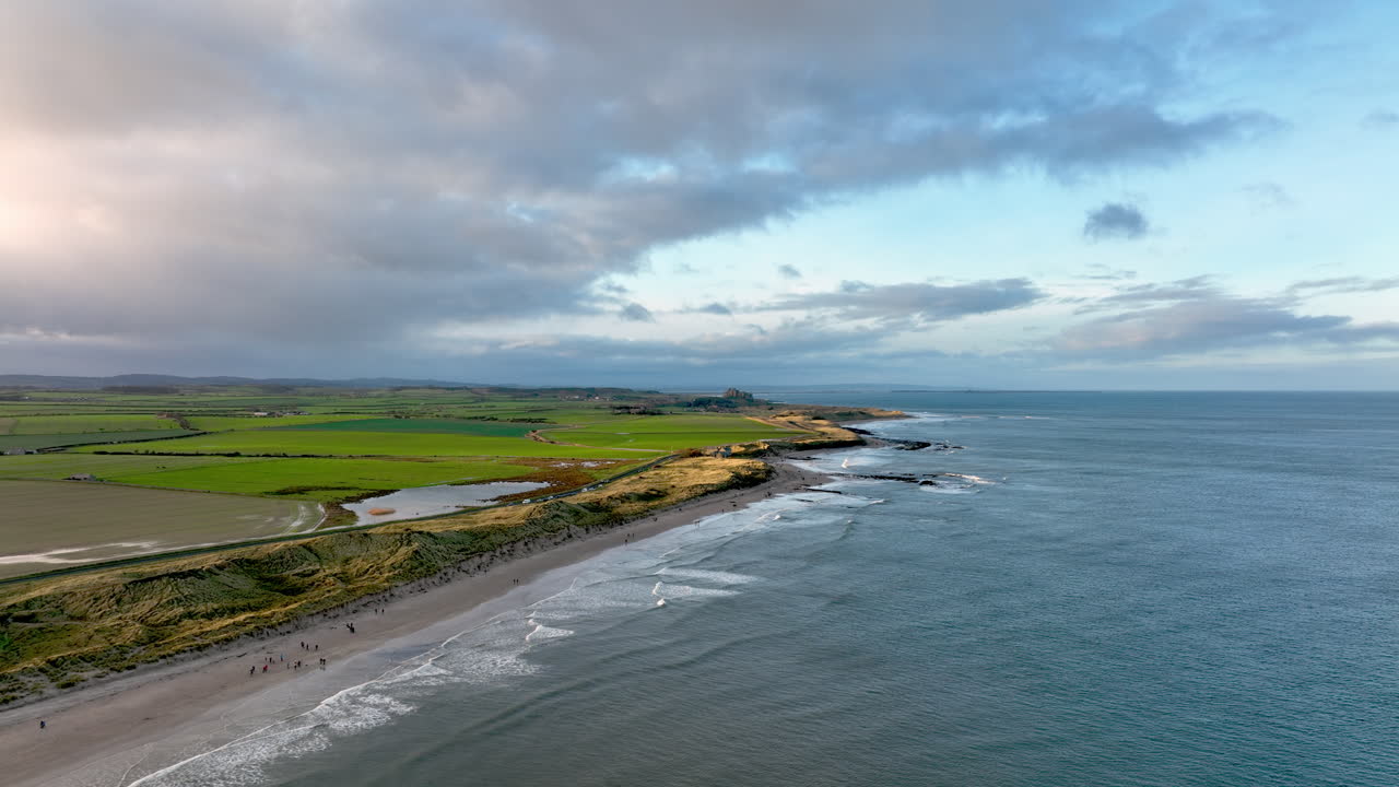 4K Flygbild över Seahouses kustlinje med Bamburgh Castle i fjärran vid solnedgången