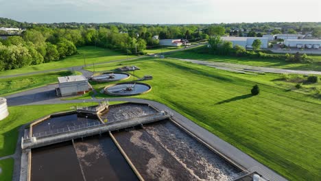wastewater treatment plant with splashing water in american factory. sunset time with green lawn and green trees in spring. aerial rising wide shot.