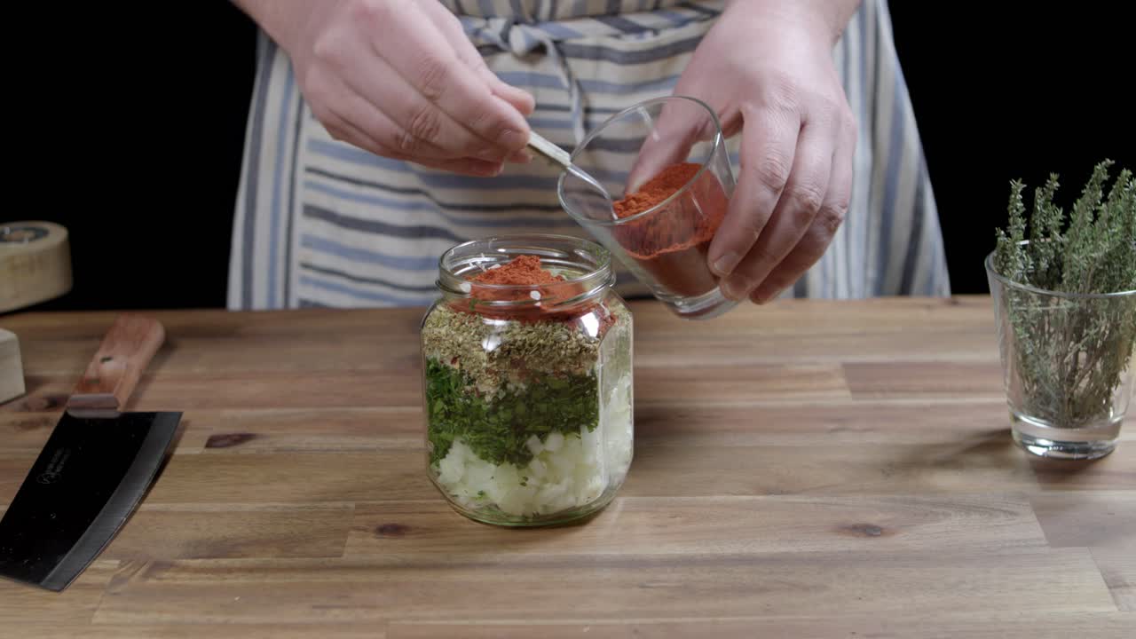 Chef adds red pepper with a spoon to chimichurri sauce mixture in transparent container, argentine cuisine