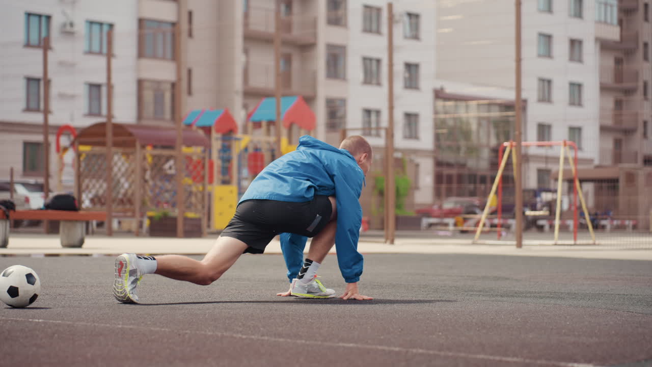 urban playground soccer warmup white male leans forward tying sneaker near soccer ball on wet asphalt, blue jacket glistening under drizzle, empty residential playground and swings in background