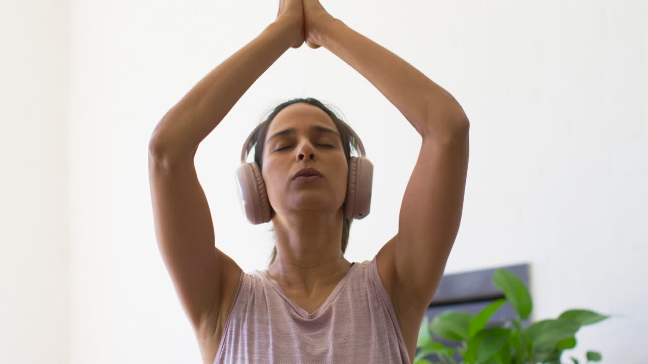 Mujer escuchando un audio de bienestar a través de auriculares, estirándose hacia arriba y luego uniendo las palmas a la altura del pecho.