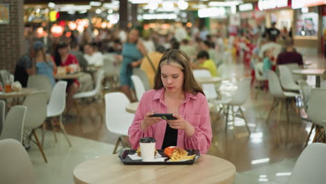 white lady in pink dress standing at a wooden table in a busy restaurant taking picture of her meal, sits down to take a picture of her meal, with background slightly blurred with people