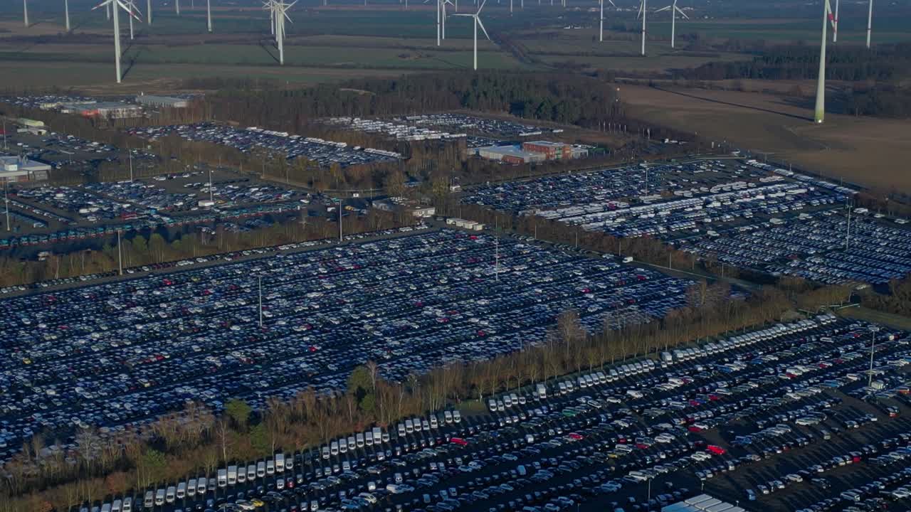 used car trade awaiting transport at a large distribution center with warehouses and railway tracks, surrounded by wind turbines. Magic aerial view ascending raise up drone