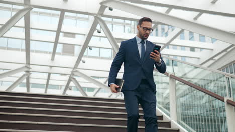 worrying caucasian male office worker rushing down the steps on his business with notebook looking at phone screen