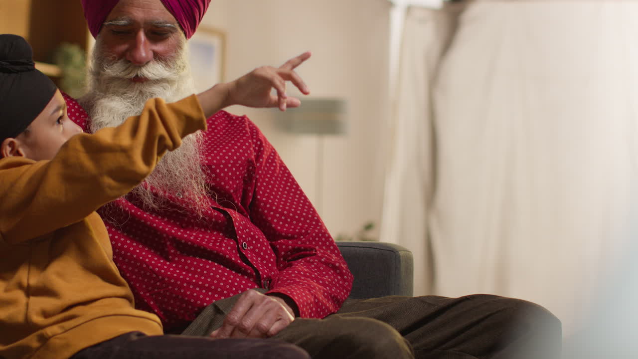 Smiling Sikh Grandfather And Grandson Wearing Turbans Sitting On Sofa At Home Talking Together 2