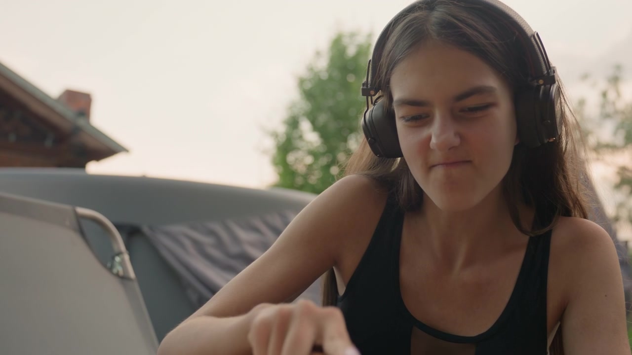 Giovane donna con le cuffie prepara il cibo a un tavolo all'aperto con tagliere e verdure. La donna affetta gli ingredienti mentre è seduta in una luce naturale soffusa, circondata da un ambiente da pranzo informale.