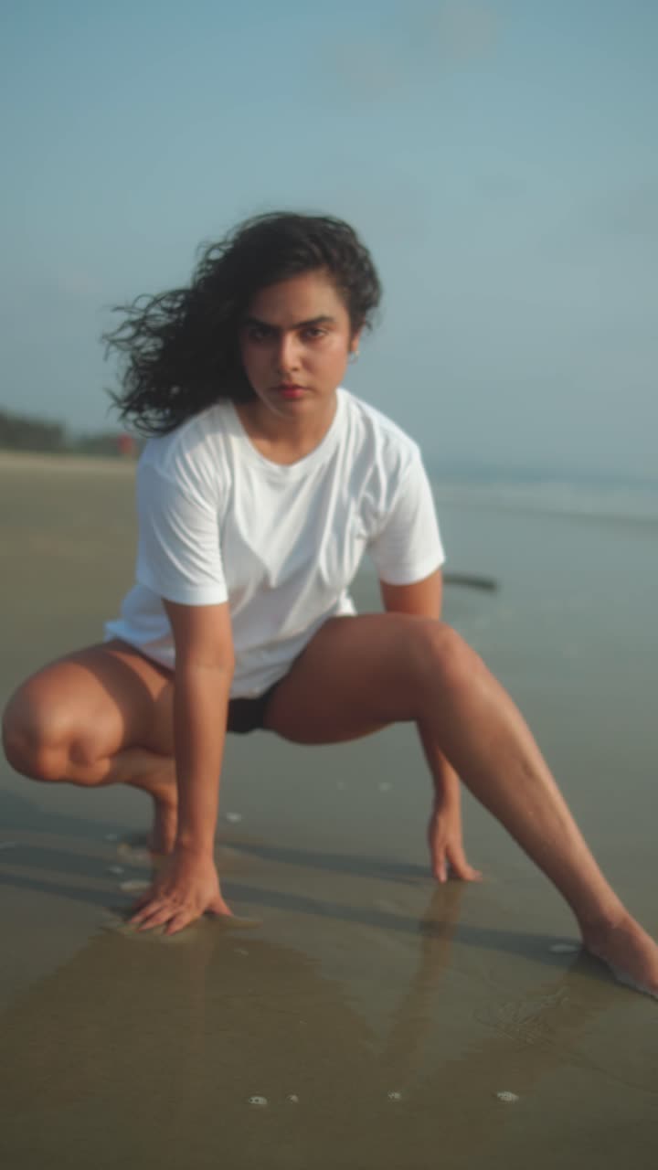 Wanita India mengenakan kaus putih dan bawahan bikini berjongkok dengan kuda-kuda lebar di pasir basah