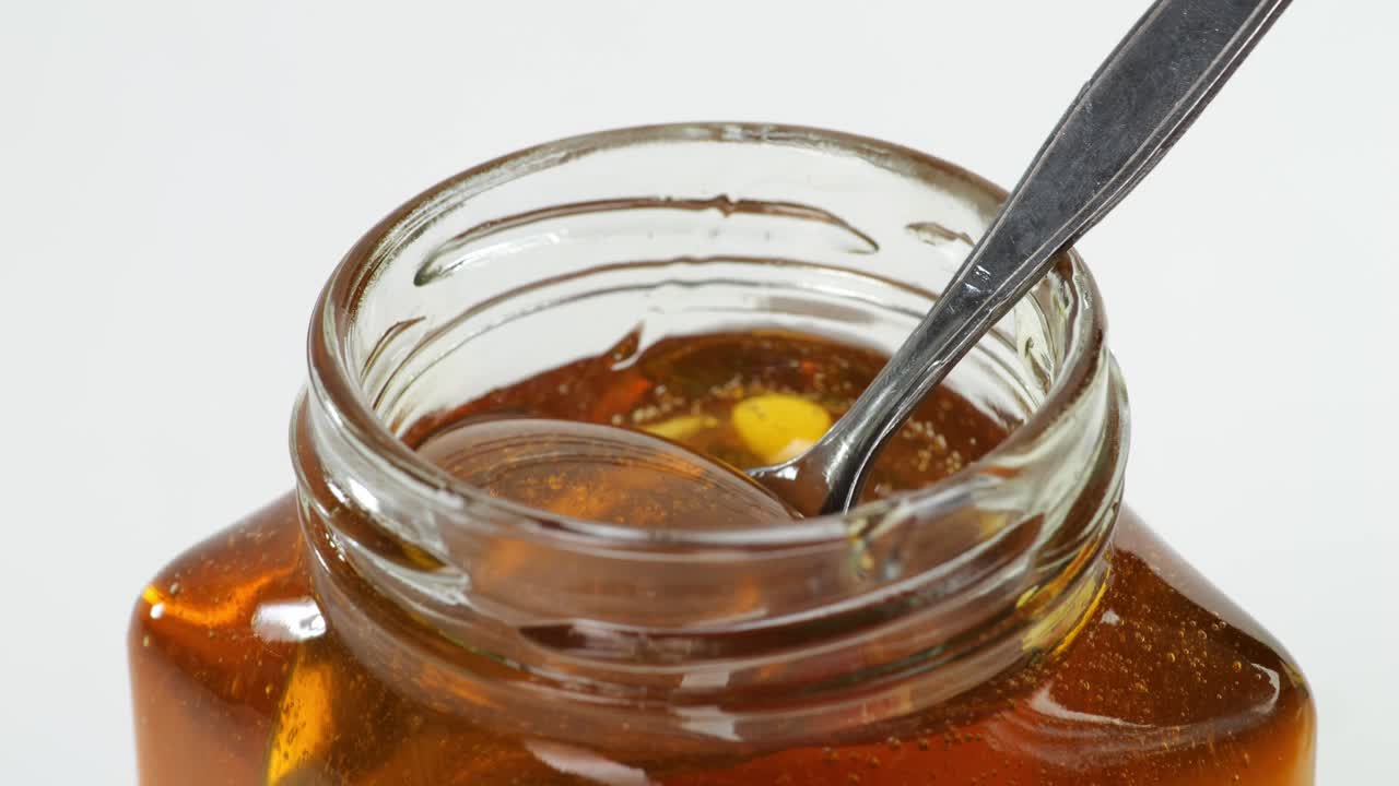A Metal Spoon Is Dipped To Take Honey In A Jar On The White Background. Honey Dripping From Metal Spoon, Healthy Organic Thick Honey Closeup, Sweet Honey Dessert 4K. Slow Motion