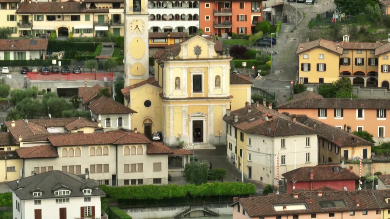 Storica Chiesa di San Giorgio Martire e colorati edifici del villaggio circondati dal verde nel Lago d'Iseo, Italia, ripresa aerea in discesa