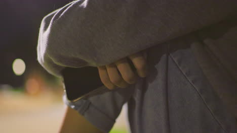 close-up of individual casually holding cell phone in hand, wearing a gray sleeve and jeans, with soft background lighting and blurred park scenery creating a calm atmosphere at night