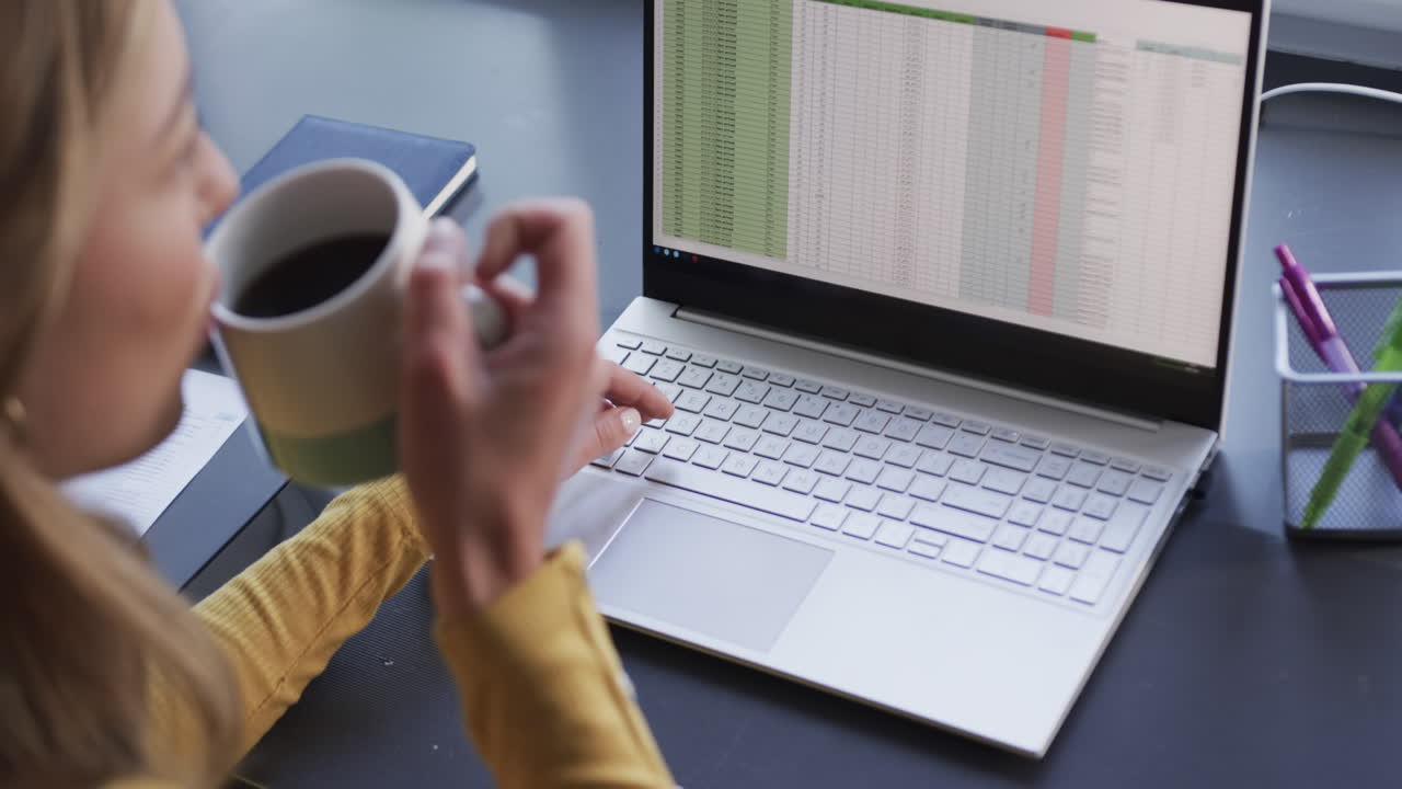 Biracial woman sitting at desk using laptop and drinking coffee at home, slow motion