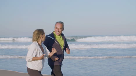 una alegre pareja madura corriendo a lo largo de la costa del océano por la mañana