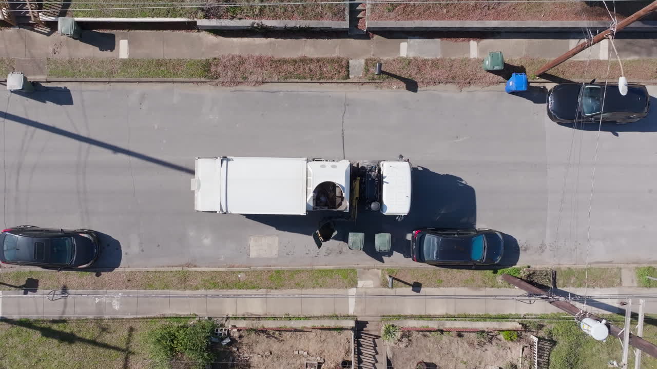 Top down aerial drone footage slowly moving down towards a dump truck that is grabbing recycling cans.