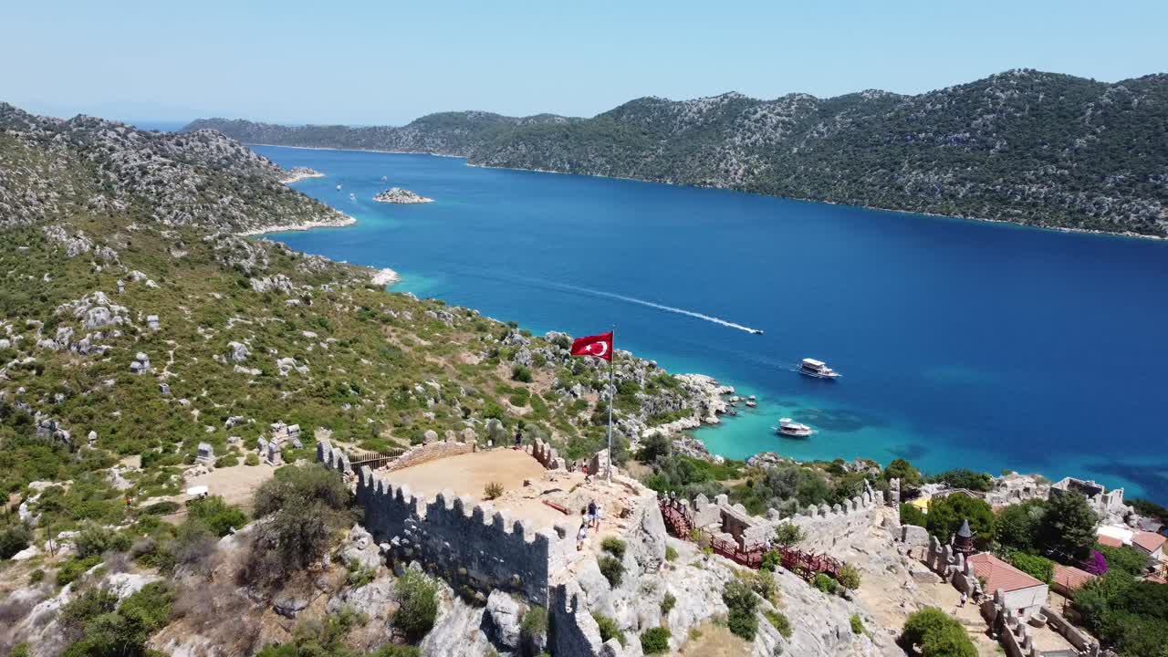 Antalya, Kaleköy'de mavi koyu gören dişli surlara sahip kaleyi ve kırmızı kiremitli köyü, engebeli tepeleri, berrak suyu, sıcak gün ışığını ve sakin kıyı atmosferini gösteren havadan drone çekimi