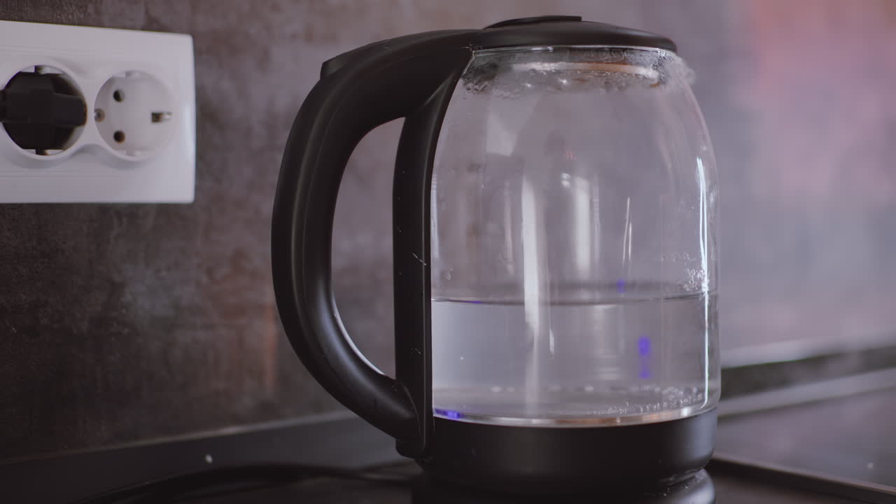 Primo piano di una mano che spegne un bollitore elettrico con corpo in vetro trasparente pieno d'acqua, manico e base neri, collegato a una presa a muro durante la routine quotidiana
