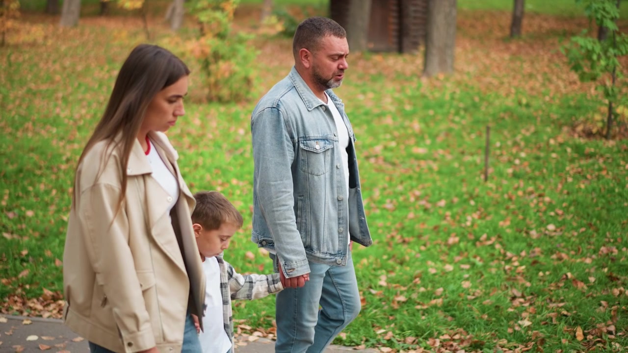 Primo piano di una famiglia che cammina insieme in un parco autunnale, con la donna e l'uomo che si tengono per mano mentre sono accompagnati da un bambino piccolo. La scena presenta erba verde cosparsa di foglie cadute, alberi che mostrano.
