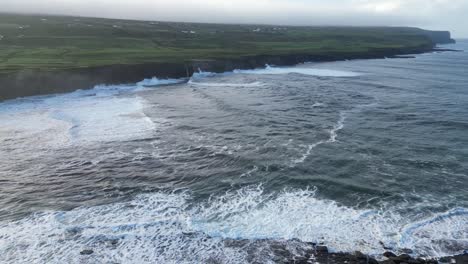 Salvajes-Olas-Del-Atlántico-Rompiendo-Contra-Los-Acantilados-De-Doolin,-Irlanda,-Con-Exuberantes-Campos-Verdes-En-El-Fondo,-Vista-Aérea