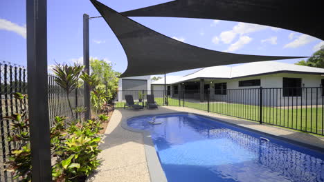 gran lujosa piscina contemporánea al aire libre en el suelo, agua azul clara valla negra cubierta de hormigón gris hogar moderno hierba verde cielo azul velas de sombra oscura área de entretenimiento al aire libre