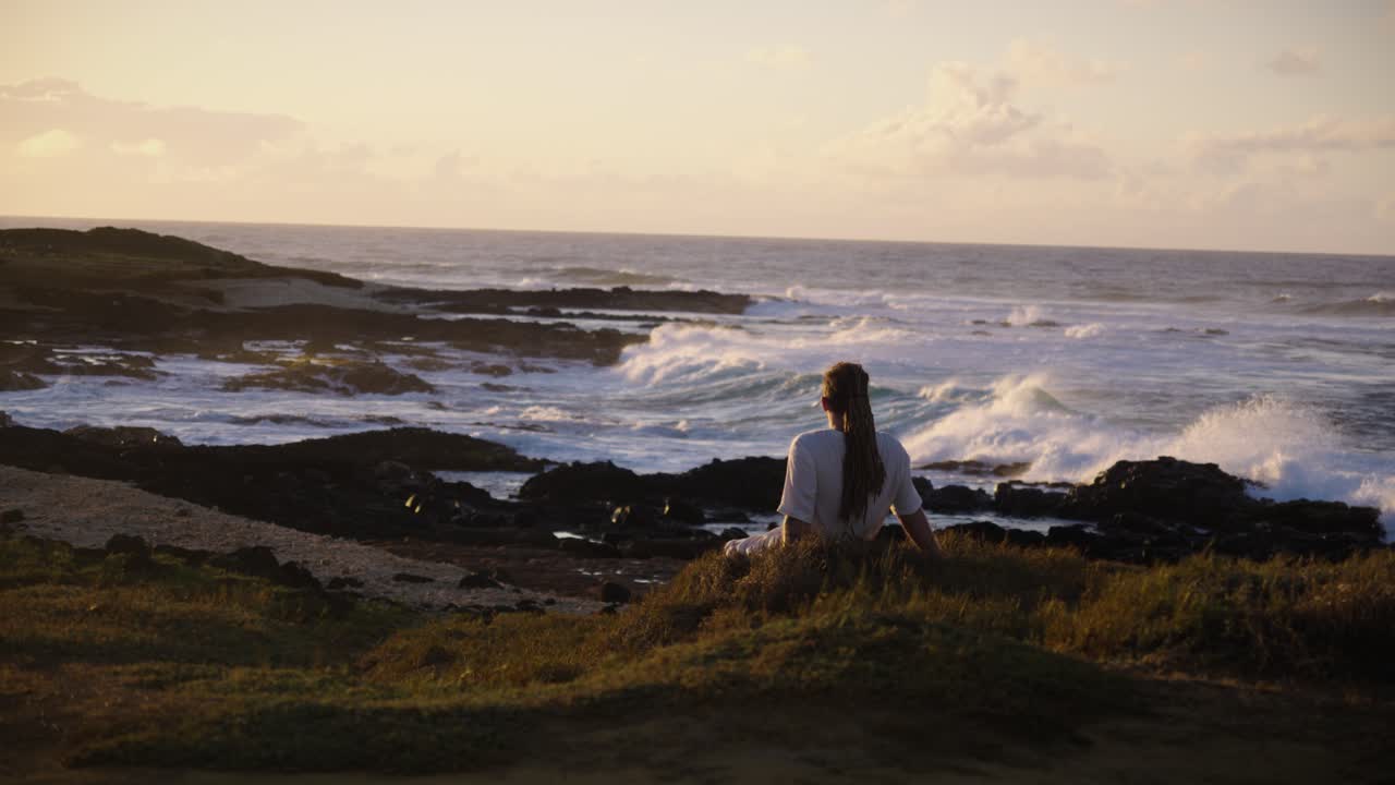 A man sits in solitude on a grassy bluff, staring at the endless ocean horizon as waves surge against the dark rocks below in golden sunset glow