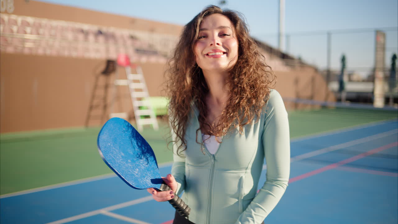 A happy and smiling woman with curly hair holding a Pickleball blue racket and a yellow ball on a blue court on a sunny day