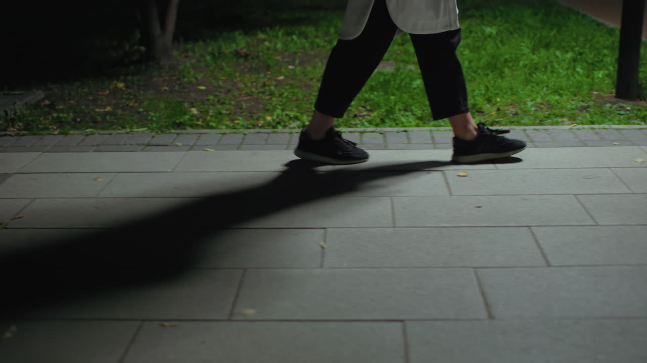 Leg view person passing bench on paved path near greenery at night, subtle light casting strong shadow, capturing calm steps in quiet outdoor environment, leaves scattered around surface