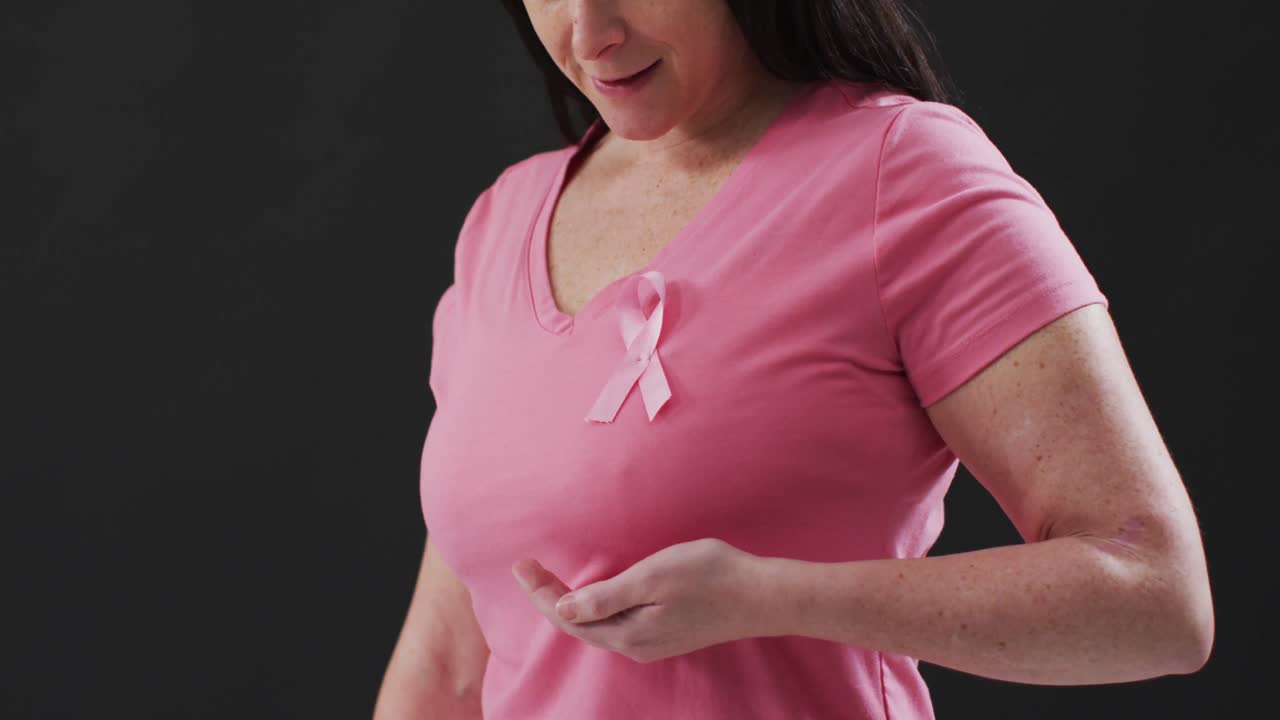 Busto di una donna che mostra il nastro rosa sul petto su sfondo nero.