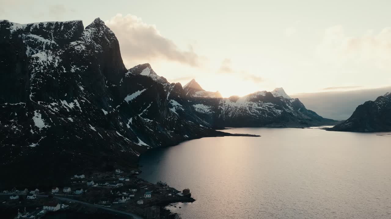 Reine, Lofoten Islands, Nordland, Norway - Snow-dusted Peaks Tower Over a Quiet Fjord as the Sun Rises Behind the Coastal Village - Orbit Drone Shot