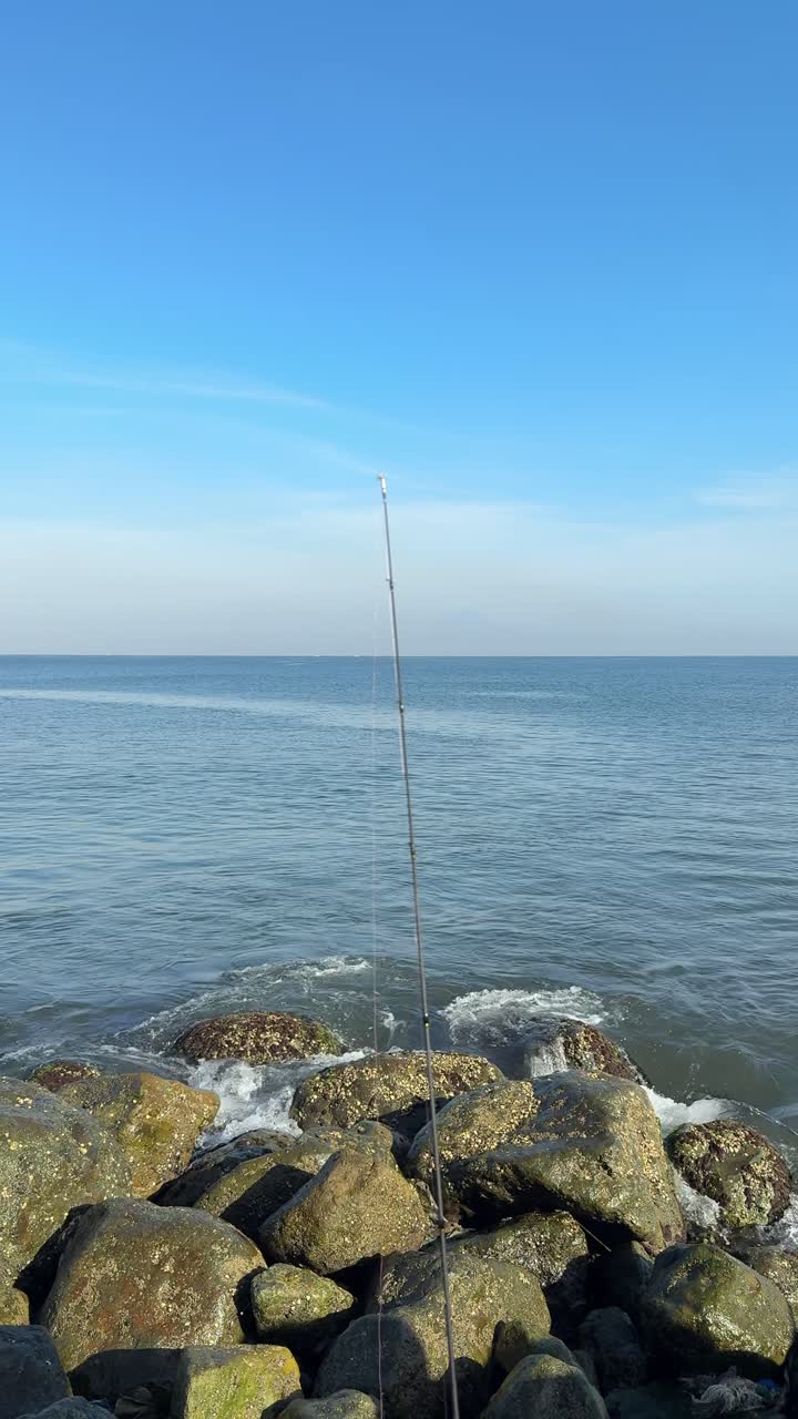 Canna da pesca su costa rocciosa con oceano e cielo azzurro.