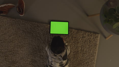 overhead shot of young boy lying on rug at home at home playing games or streaming onto green screen digital tablet at night