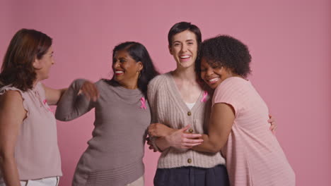 retrato de estudio de un grupo multirracial de mujeres sonrientes de diferentes edades que llevan ropa rosa y cintas de concienciación sobre el cáncer de mama abrazándose contra un fondo rosa 1