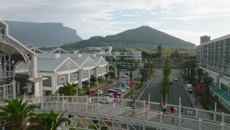 Forwards-fly-above-wide-street-with-tropical-trees.-People-walking-on-footbridge-connecting-buildings-of-Victoria-Wharf-Shopping-Centre.-City-and-mountains-in-background.-Cape-Town,-South-Africa