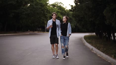 stylish young people are walking by green park with trees and talking. wearing casual trendy clothes. front view, slow motion