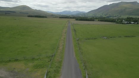 Countryside-road-running-between-green-fields-with-mountains-on-horizon-on-cloudy-summer-day