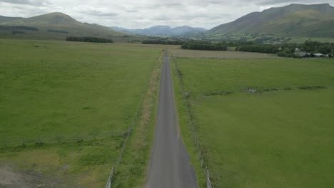 countryside road running between green fields with mountains on horizon on cloudy summer day