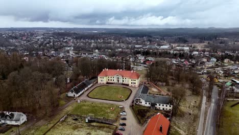 Aerial-drone-of-Zante-Manor,-a-stately-yellow-and-red-manor-house,-sits-amidst-a-sprawling-complex-of-outbuildings,-gardens,-and-grounds,-overlooking-a-quaint-town-nestled-between-rolling-hills