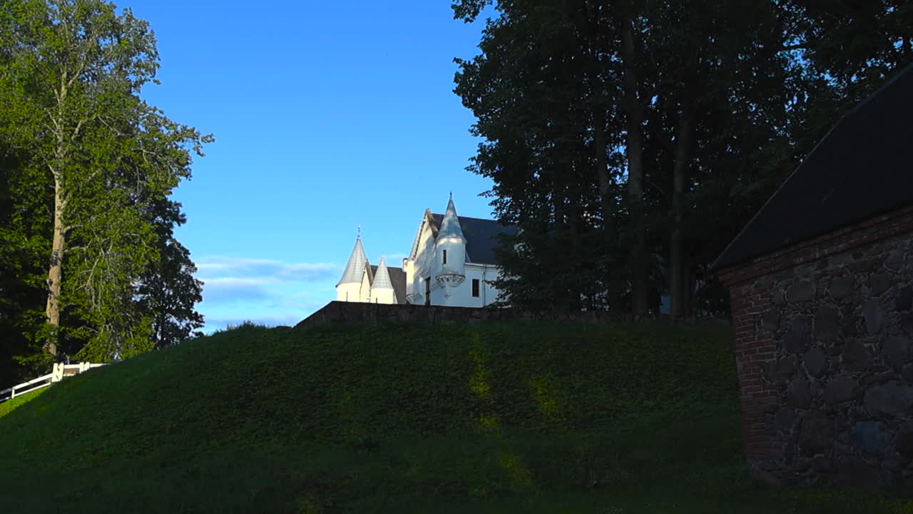 Bottom up low angle wide shot of majestic and historic white colored Alatskivi castle during summer time sunny day with forest trees around it and blue sky in the background. Castle on a hill